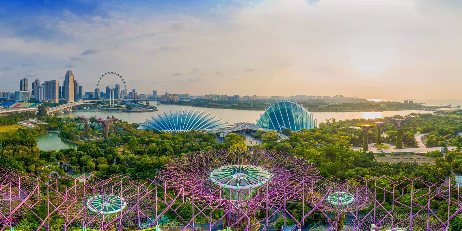 Panoramic view of Gardens by the Bay in Singapore, featuring the iconic Supertree Grove in the foreground, with the Flower Dome and Cloud Forest conservatories at the centre. The Singapore Flyer and city skyline are visible on the left, with Marina Bay and the setting sun in the background.