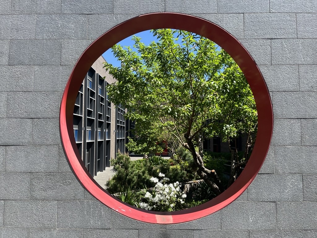 Circular, red-framed window looking onto a sunny courtyard with green trees and shrubs.