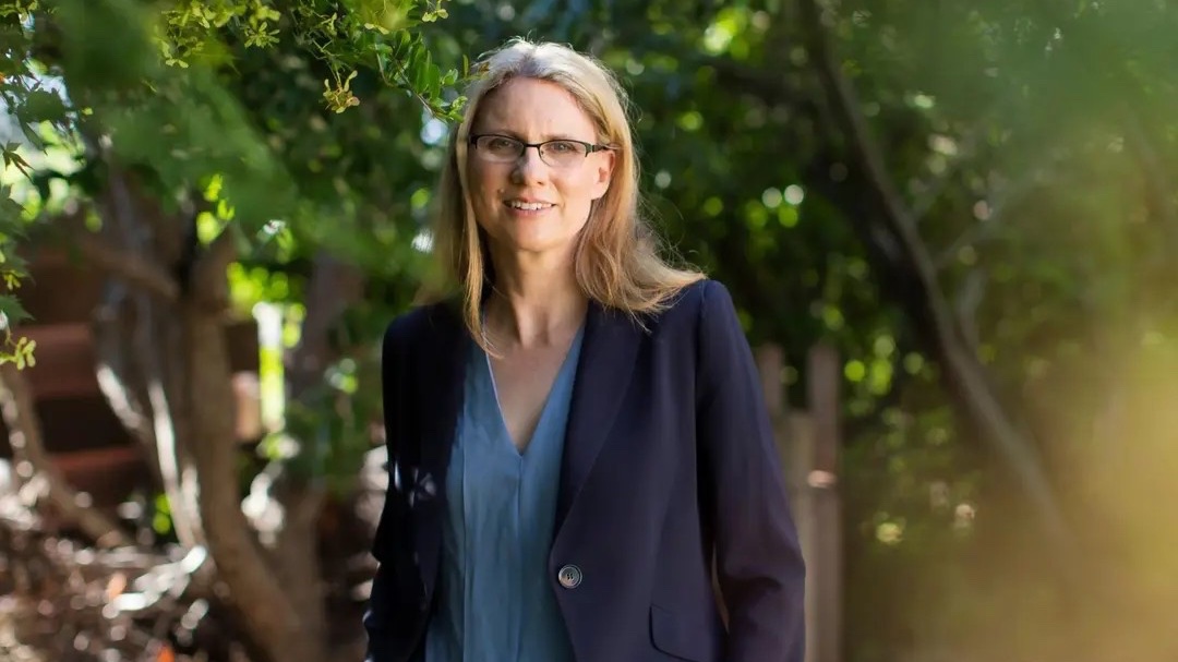 Associate Professor Lucy Neave in a blazer stands outdoors among green trees. 