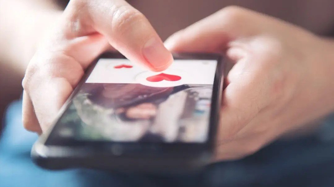Hand tapping a heart icon on a dating app profile on a smartphone.