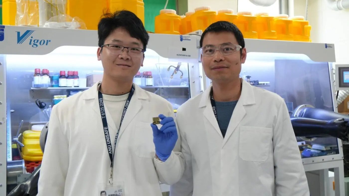 Two researchers in lab coats stand in a laboratory, one holding a small solar cell sample.