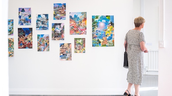 A woman walks through a bright gallery space, passing a wall of colourful collage artworks. 