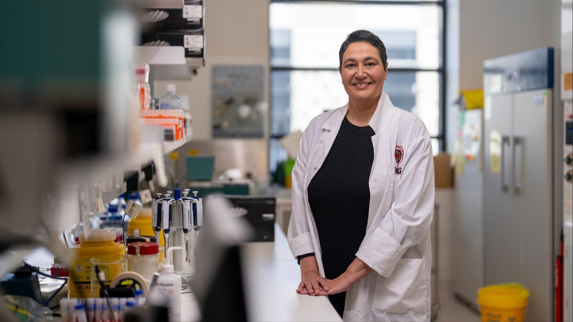 Associate Professor Azure Hermes in a white lab coat smiles while standing at a lab bench surrounded by equipment, with bright natural light from a large window behind them.