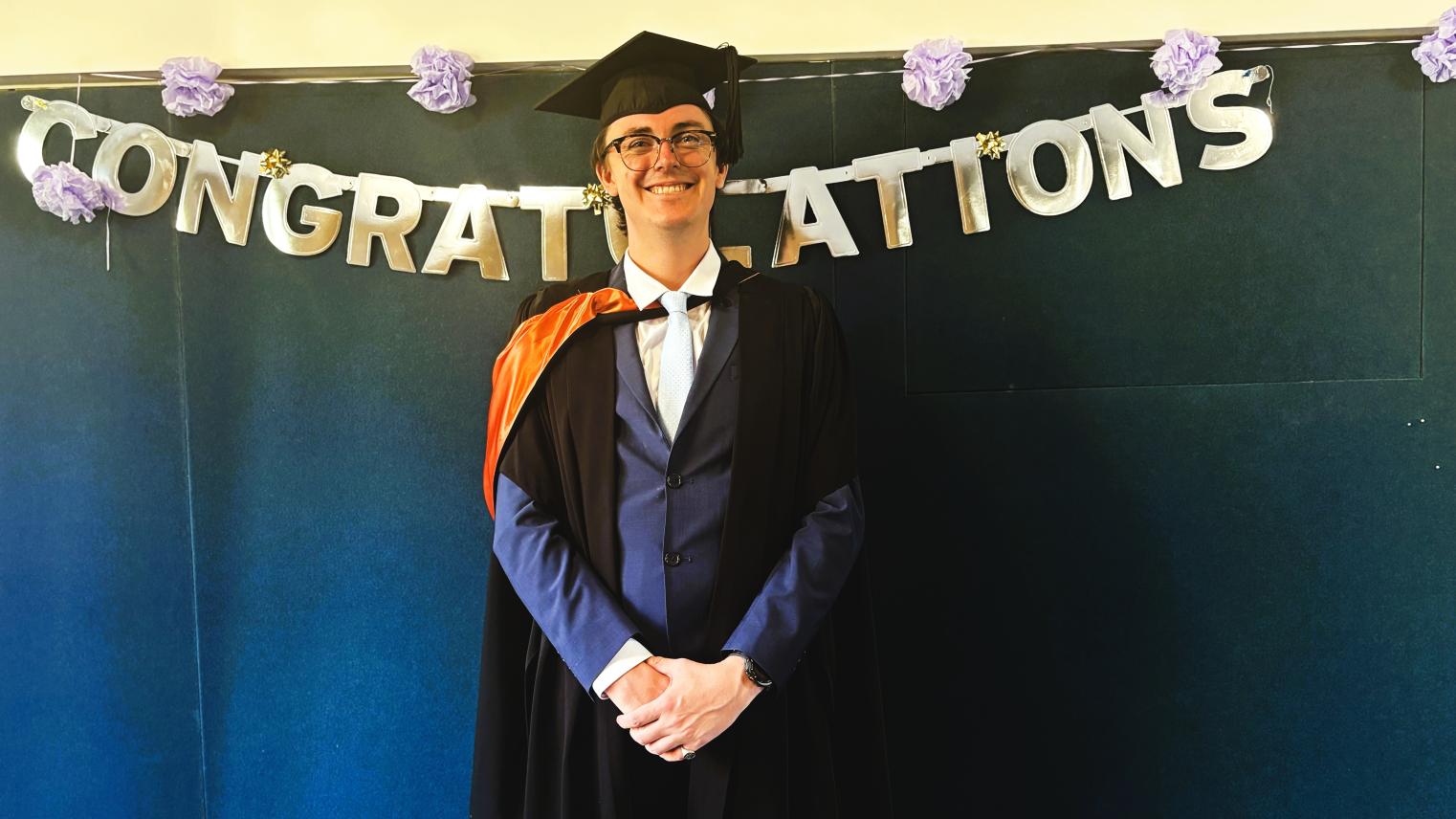 Graduate in cap and gown standing in front of a &ldquo;Congratulations&rdquo; banner.