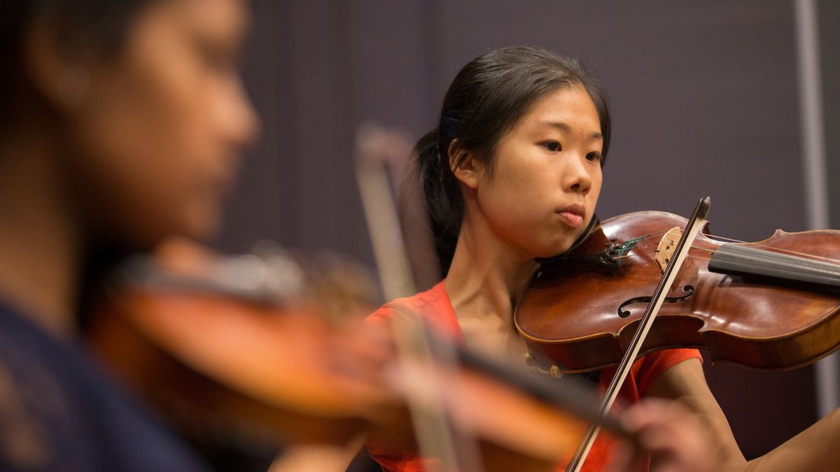 Student playing the violin during a rehearsal, with another violinist in the foreground.