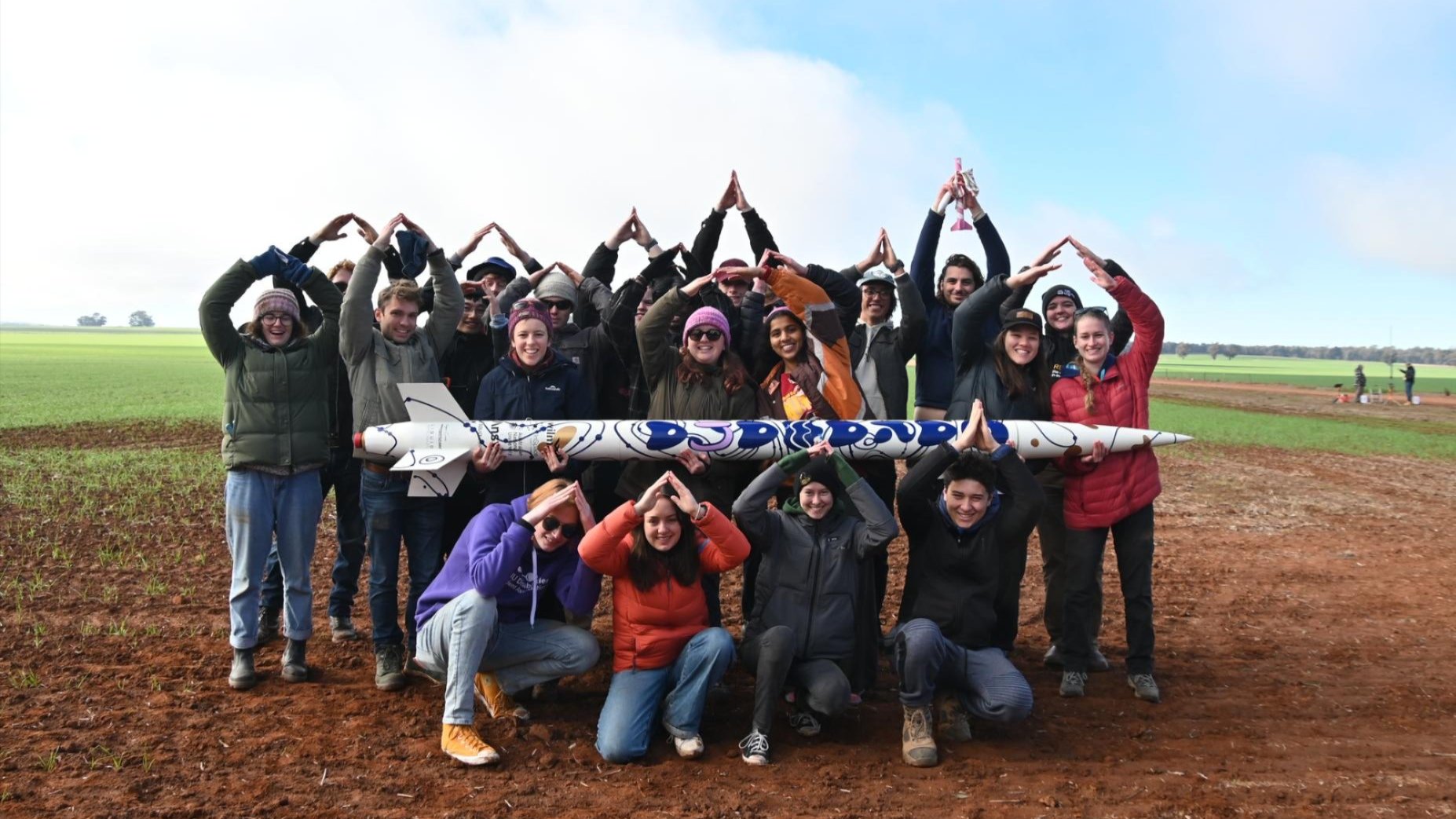 Group of students posing in a field, holding a decorated model rocket and forming triangle shapes with their arms. 