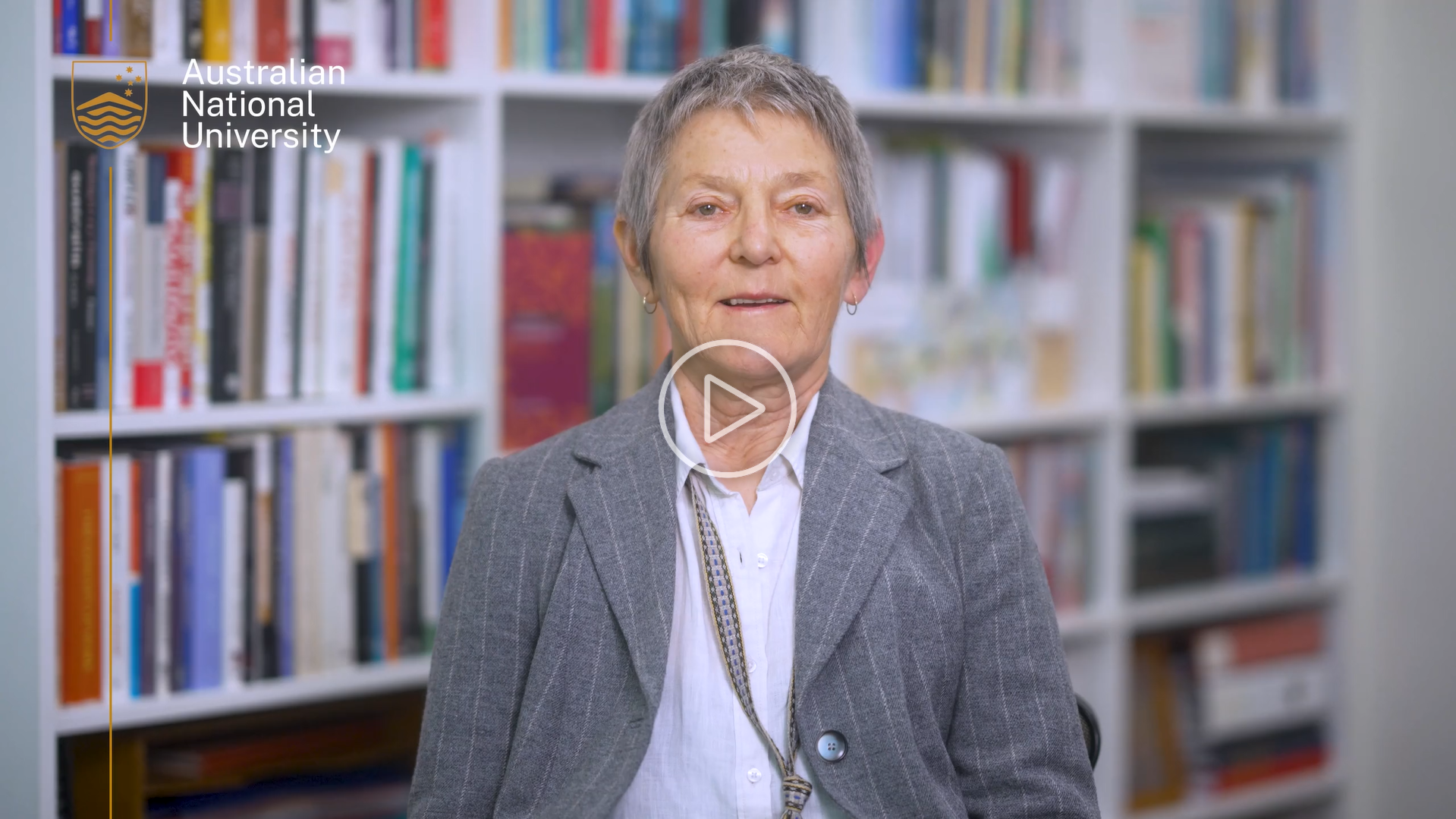 A woman in a grey blazer sits in front of bookshelves, speaking to camera with the Australian National University logo in the corner. and a play button centred on the image.
