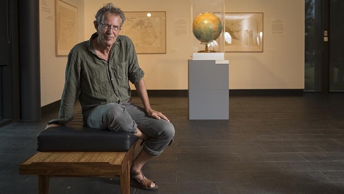 Man in a casual green shirt and grey pants sits on a wooden bench in an art gallery, with a large globe displayed behind him.