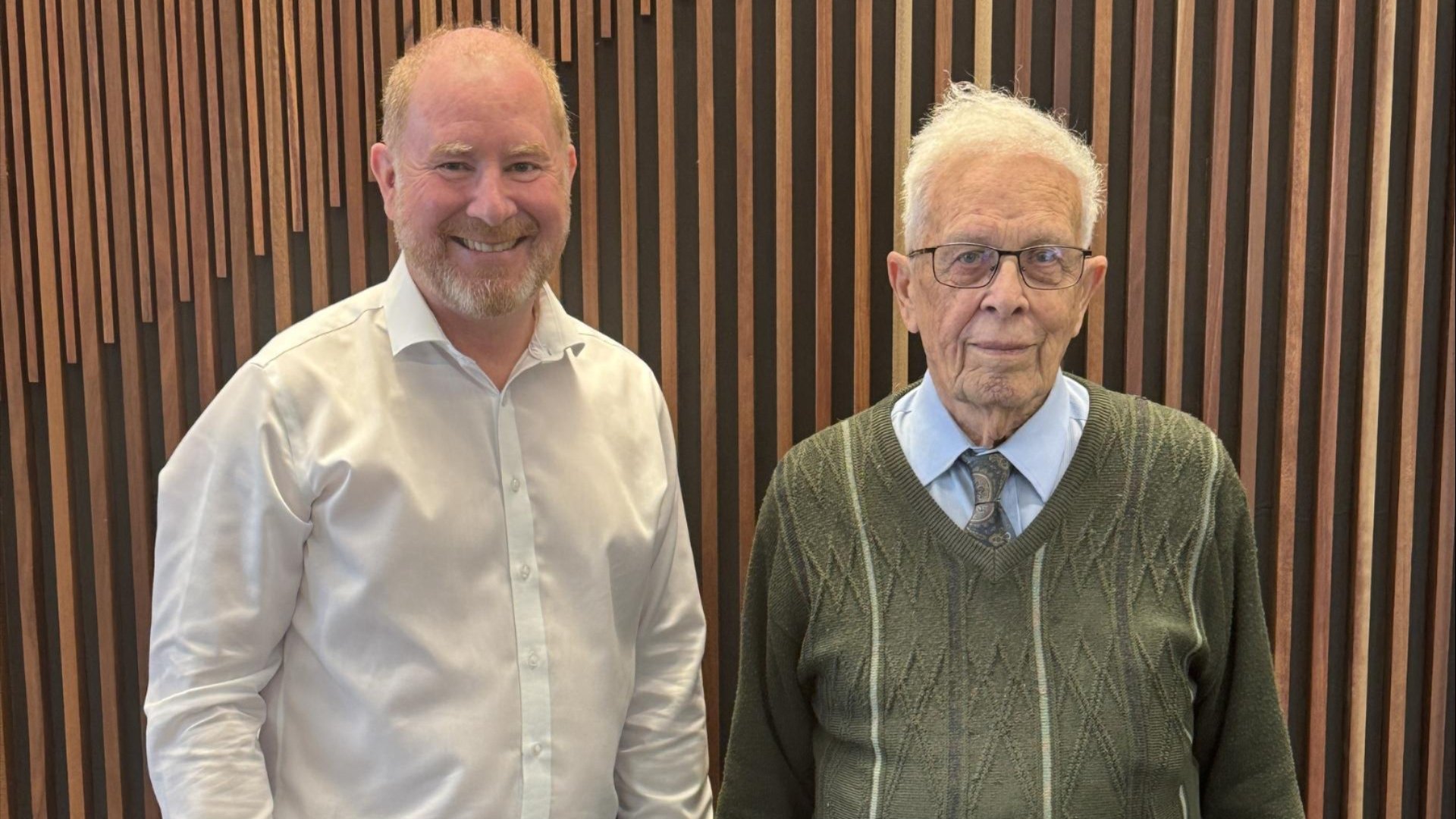 Two men standing side by side indoors against a timber wall.
