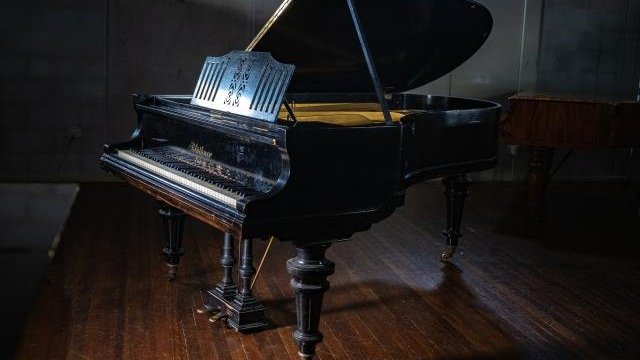 A black grand piano with an open lid sits on a polished wooden floor in a dimly lit room, light highlights the pianos glossy surface.