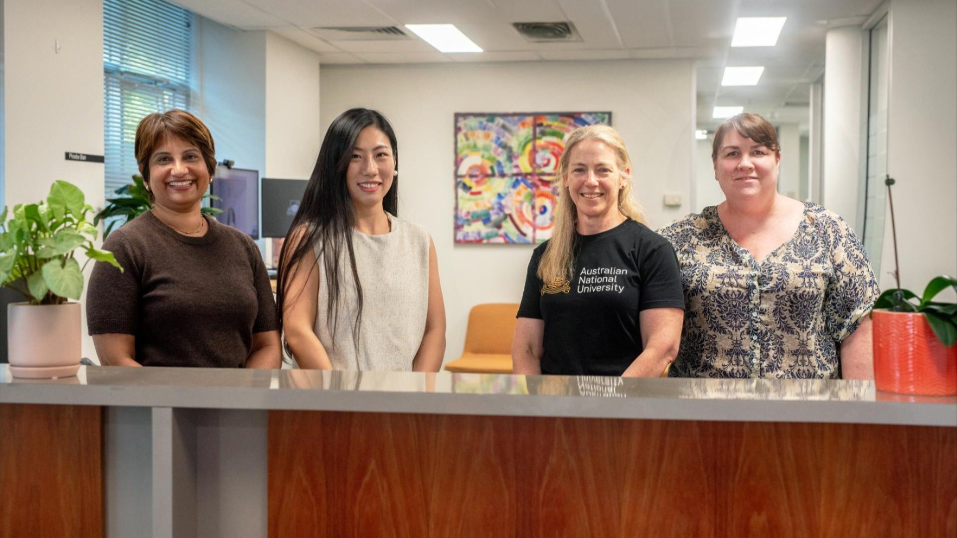 Four staff members standing behind a reception counter in an office, smiling at the camera, with plants and artwork in the background.