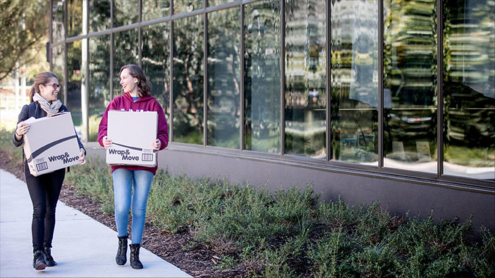 Two students walking along campus carrying moving boxes outside a glass-fronted building.