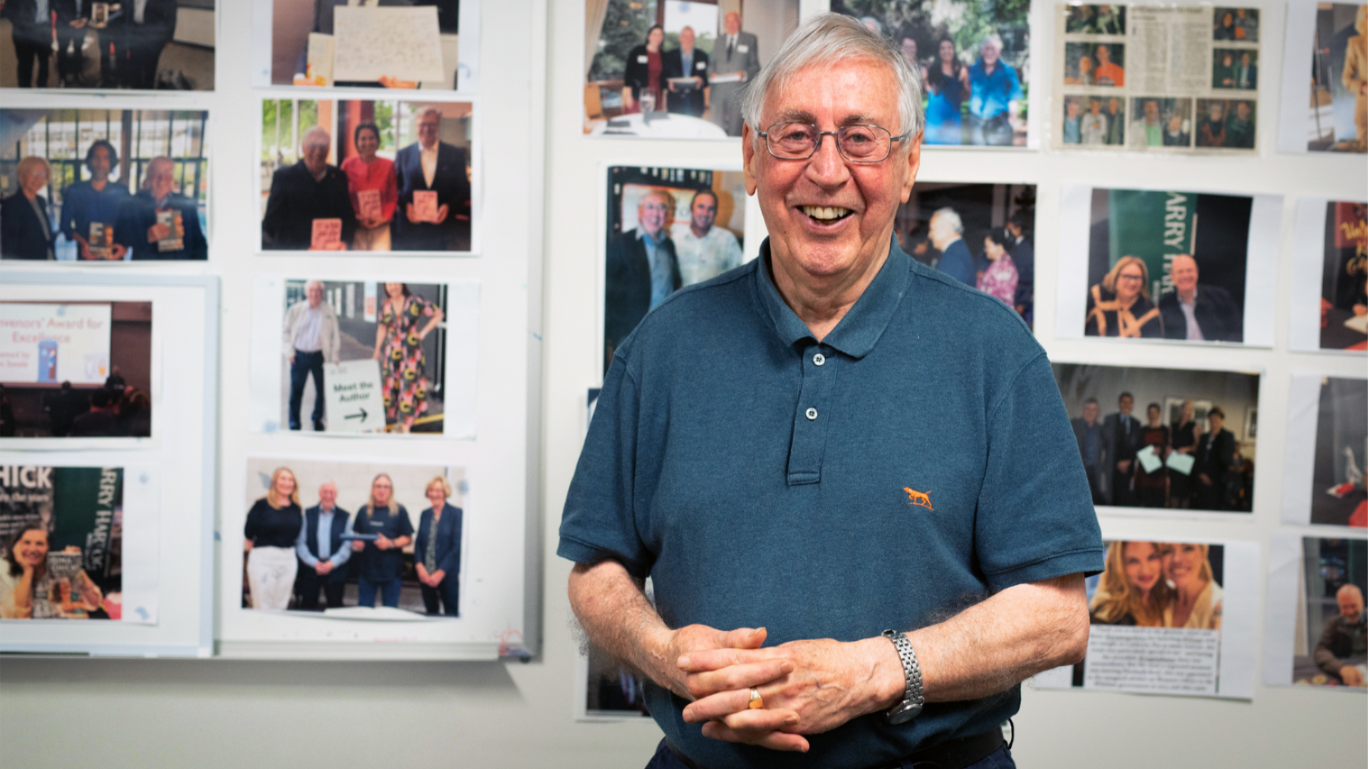 Emeritus Fellow, Colin Steele, smiling while standing in front of a wall covered with photos of events, people and achievements.