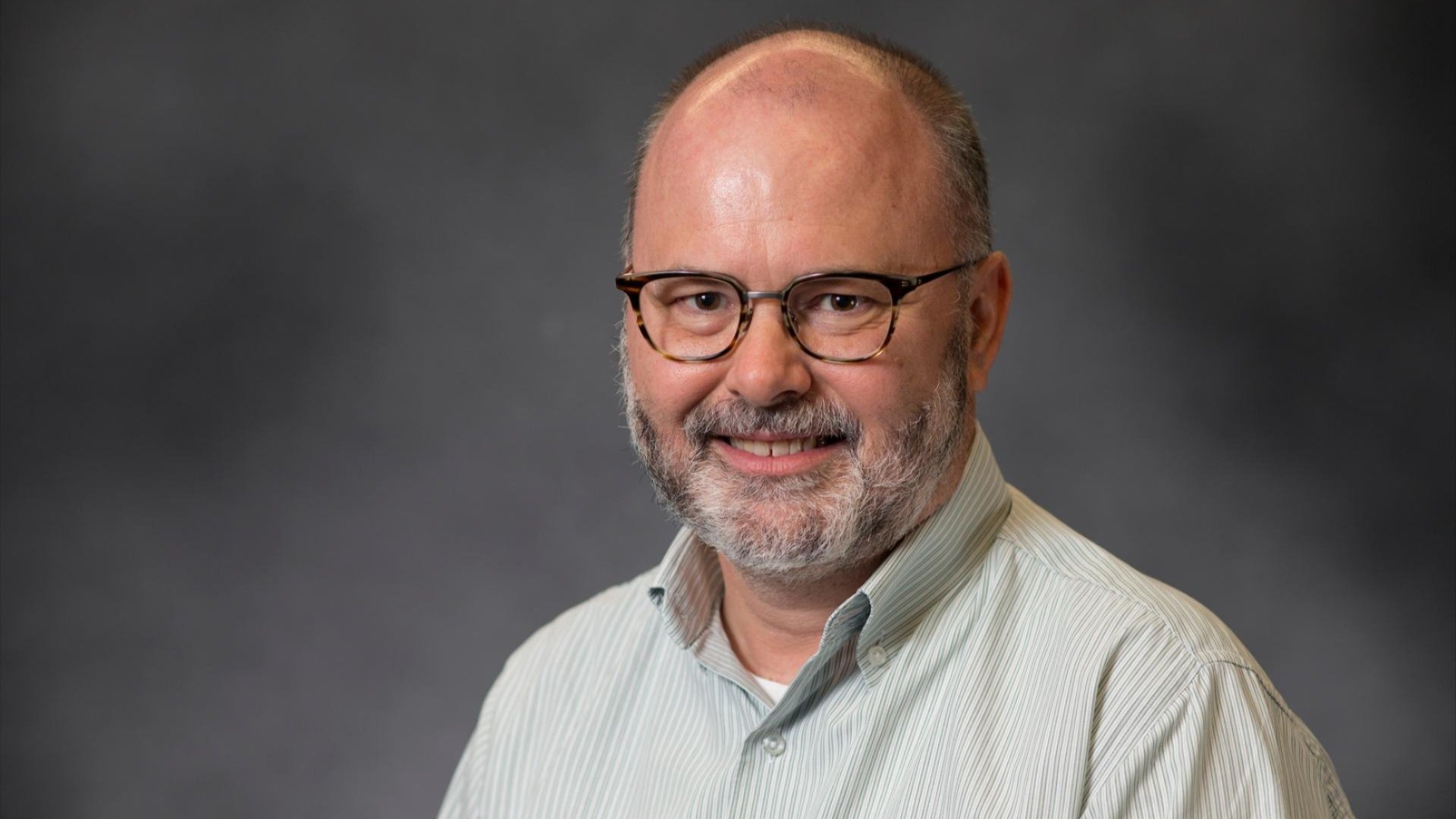 Studio portrait of Professor Royston Gustavson wearing glasses and a light green striped shirt, smiling against a grey background.