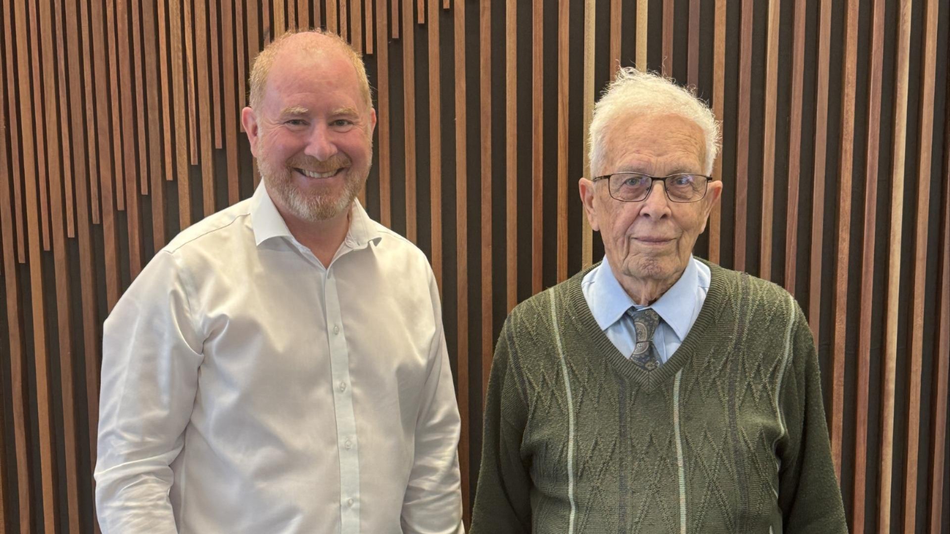 Two men standing side by side indoors against a timber wall.