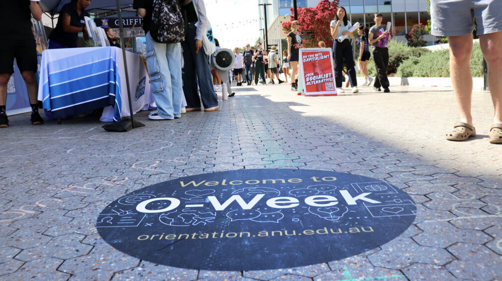 Circular O-Week welcome decal on a paved walkway, with students and stalls lining the path on campus.