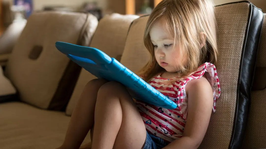 Young child sitting on a couch, focused on a tablet with a blue case.