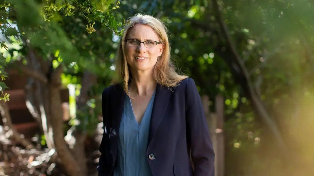 A woman in a blazer stands outdoors among green trees.