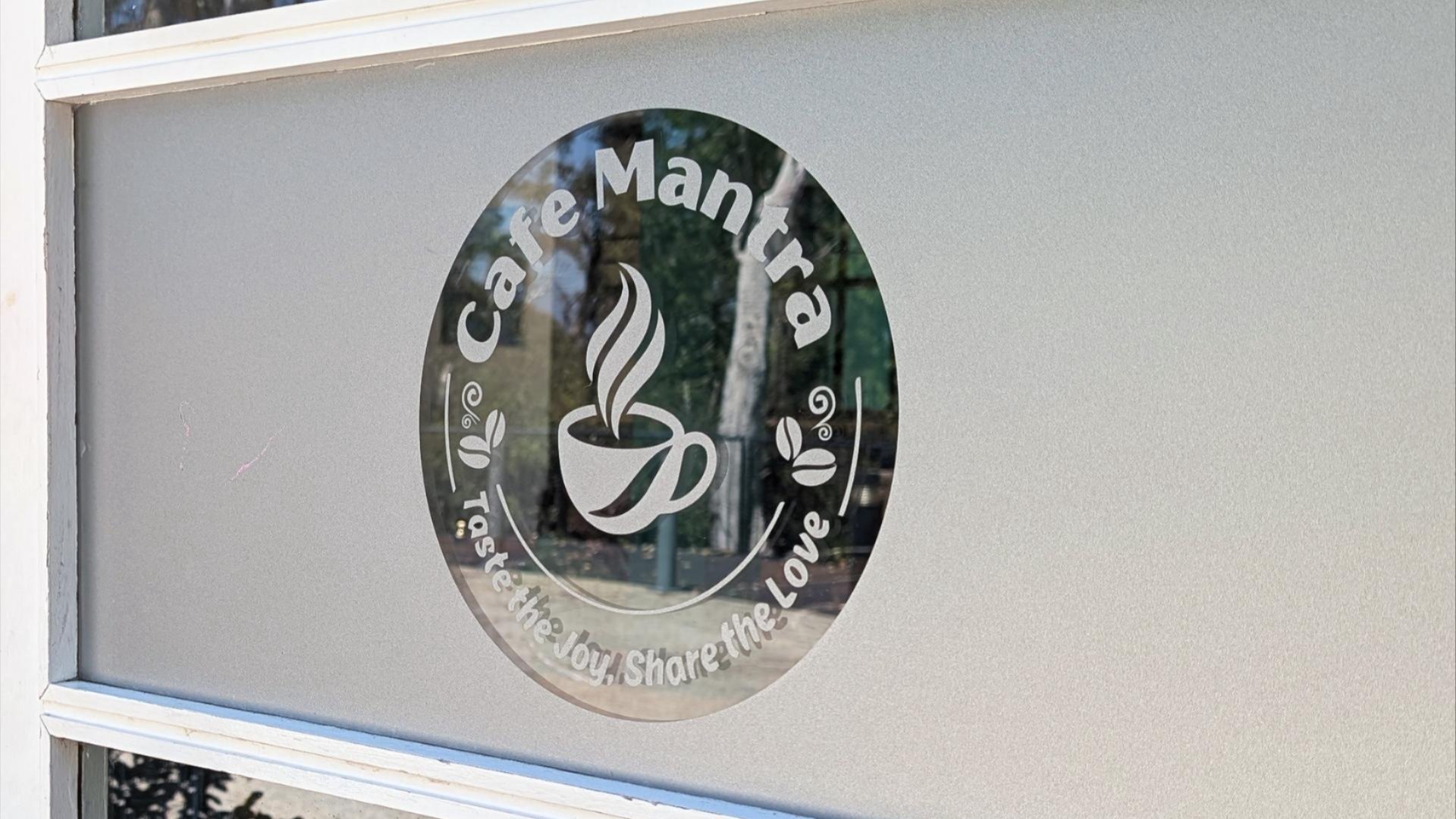 Circular &ldquo;Caf&eacute; Mantra&rdquo; logo decal on a frosted glass window, featuring a steaming coffee cup and the words &ldquo;Taste the love, share the love.&rdquo;