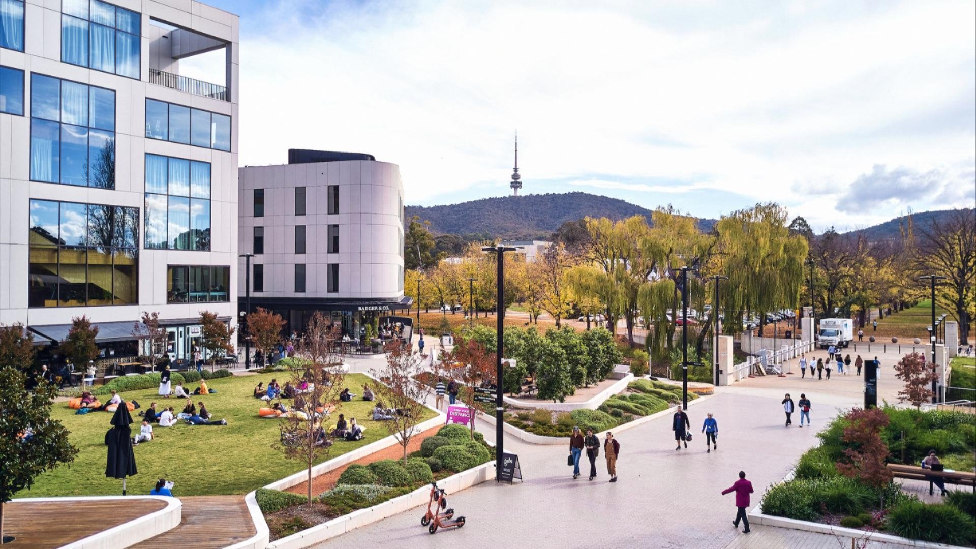 People relaxing on the lawn at Kambri, ANU, with modern buildings and Telstra Tower in the background.