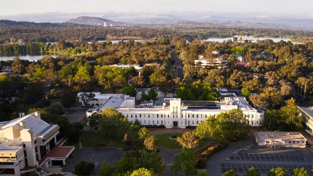 Aerial view of the Australian National University campus with the ANU School of Art and Design building centred, surrounded by trees, with Lake Burley Griffin and Canberra&rsquo;s hills visible in the background.