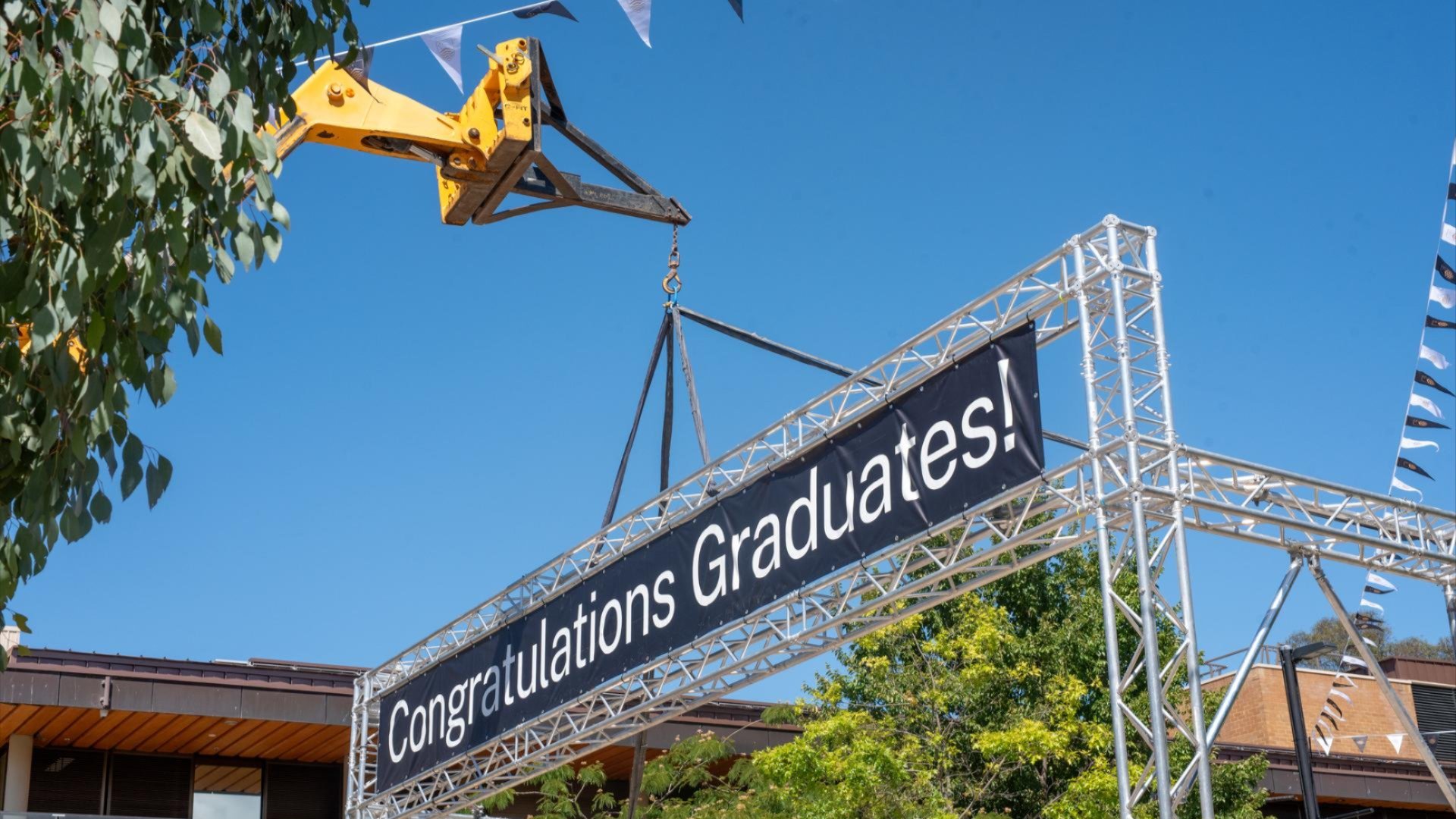 ANU workers use a crane to install a &ldquo;Congratulations Graduates!&rdquo; banner over a campus road.