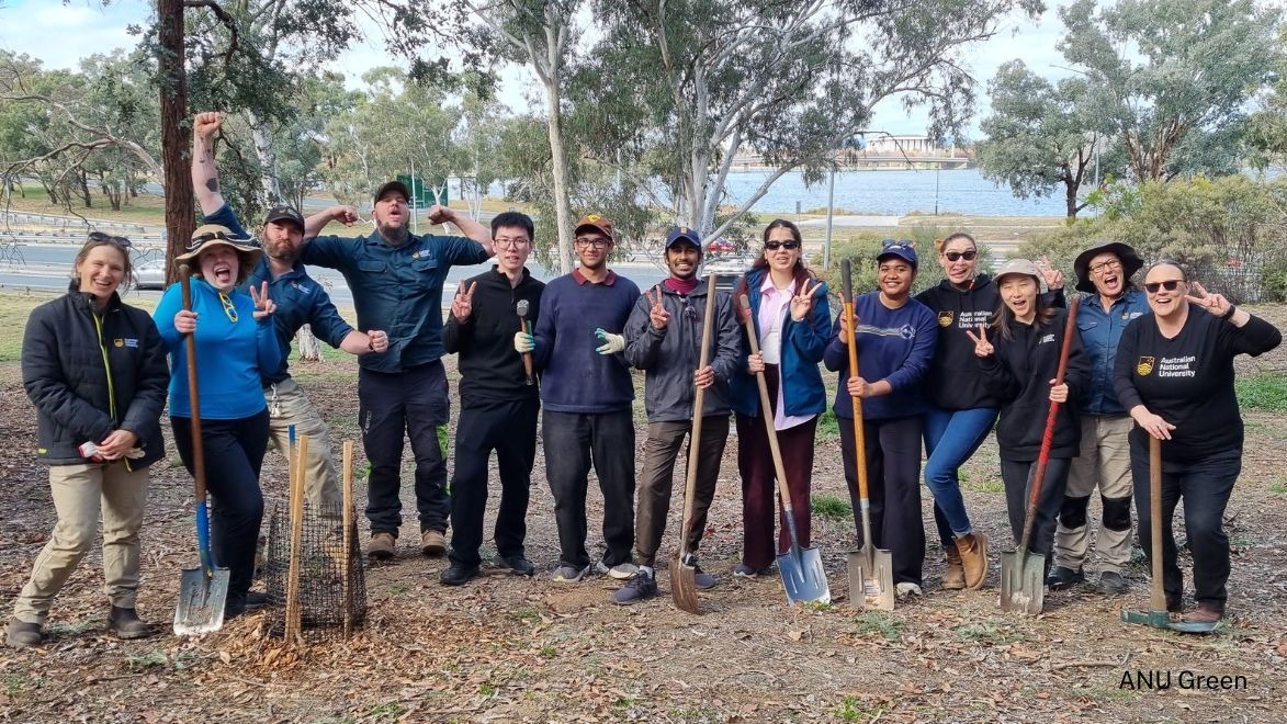 Group of students and staff smiling and posing during a campus sustainability activity, standing around a newly planted garden bed.