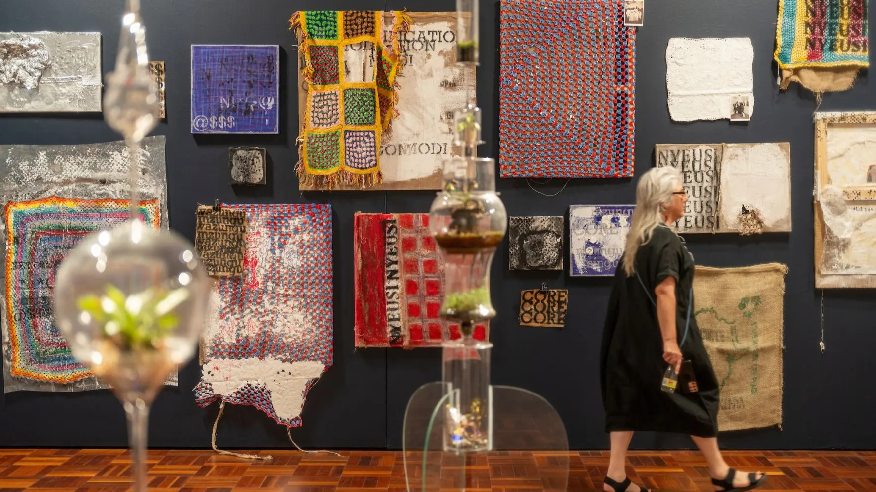 A woman walks through a gallery space showcasing painting and textile based contemporary artwork and interactive glass sculptures showcased in the 2025 Art and Design Grad Show.