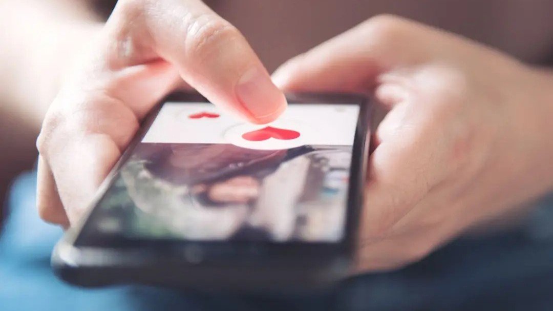 Hand tapping a heart icon on a dating app profile on a smartphone.