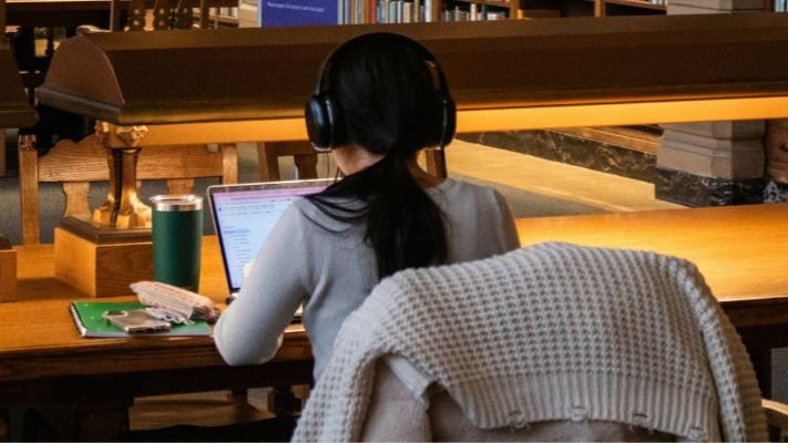 Student turned away from the camera, studying at a wooden desk in a library, wearing headphones and working on a laptop with books and a drink beside them.