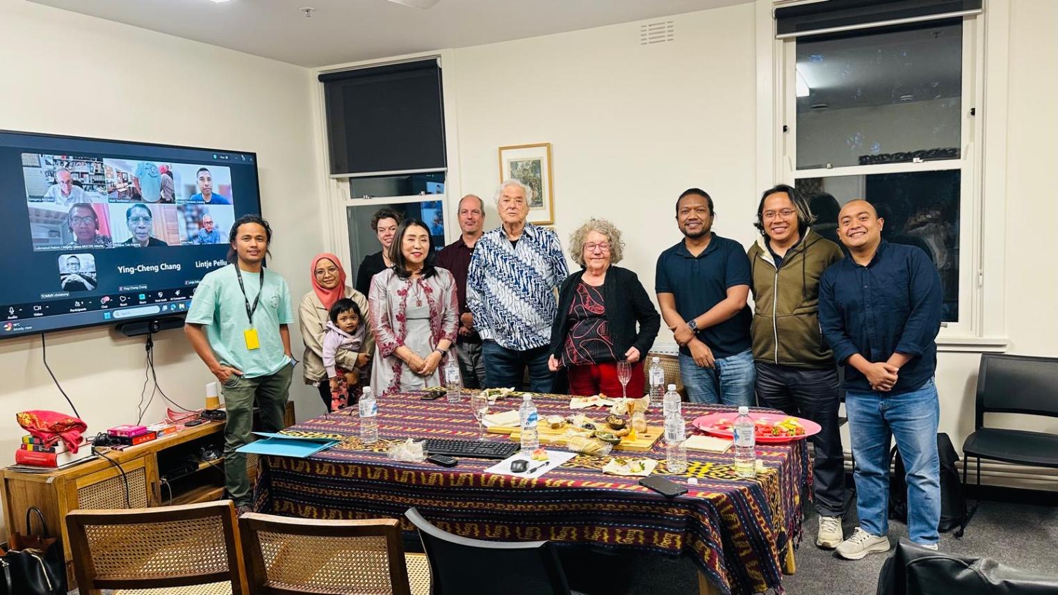 A group of people stand together in a meeting room, with others joining via video call on a screen.