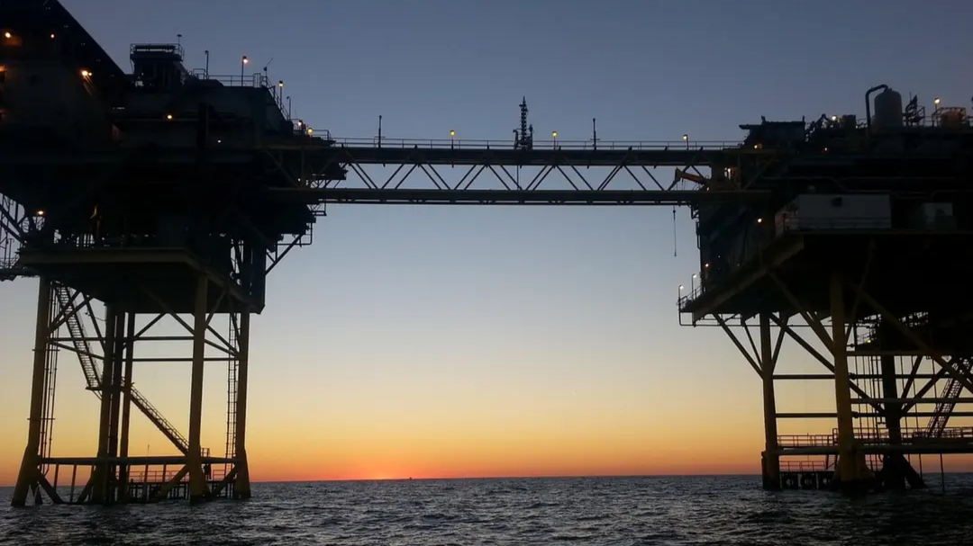 Offshore oil platform silhouetted against a sunset sky, with ocean in the foreground and the sun dipping below the horizon.