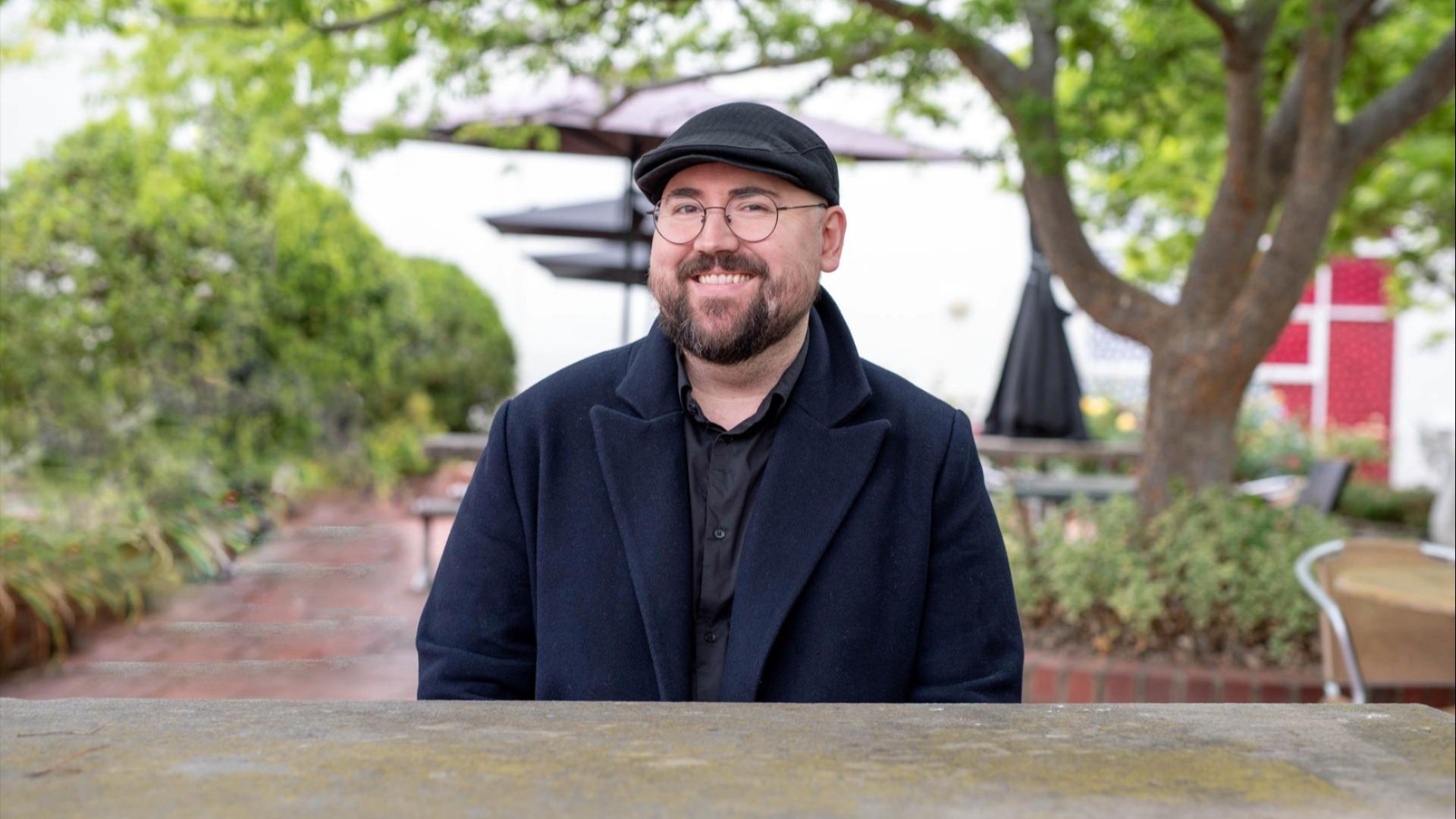 Alexander Longmore smiling while seated outdoors at a table, wearing a dark coat, black shirt, and cap, with trees and umbrellas in the background.
