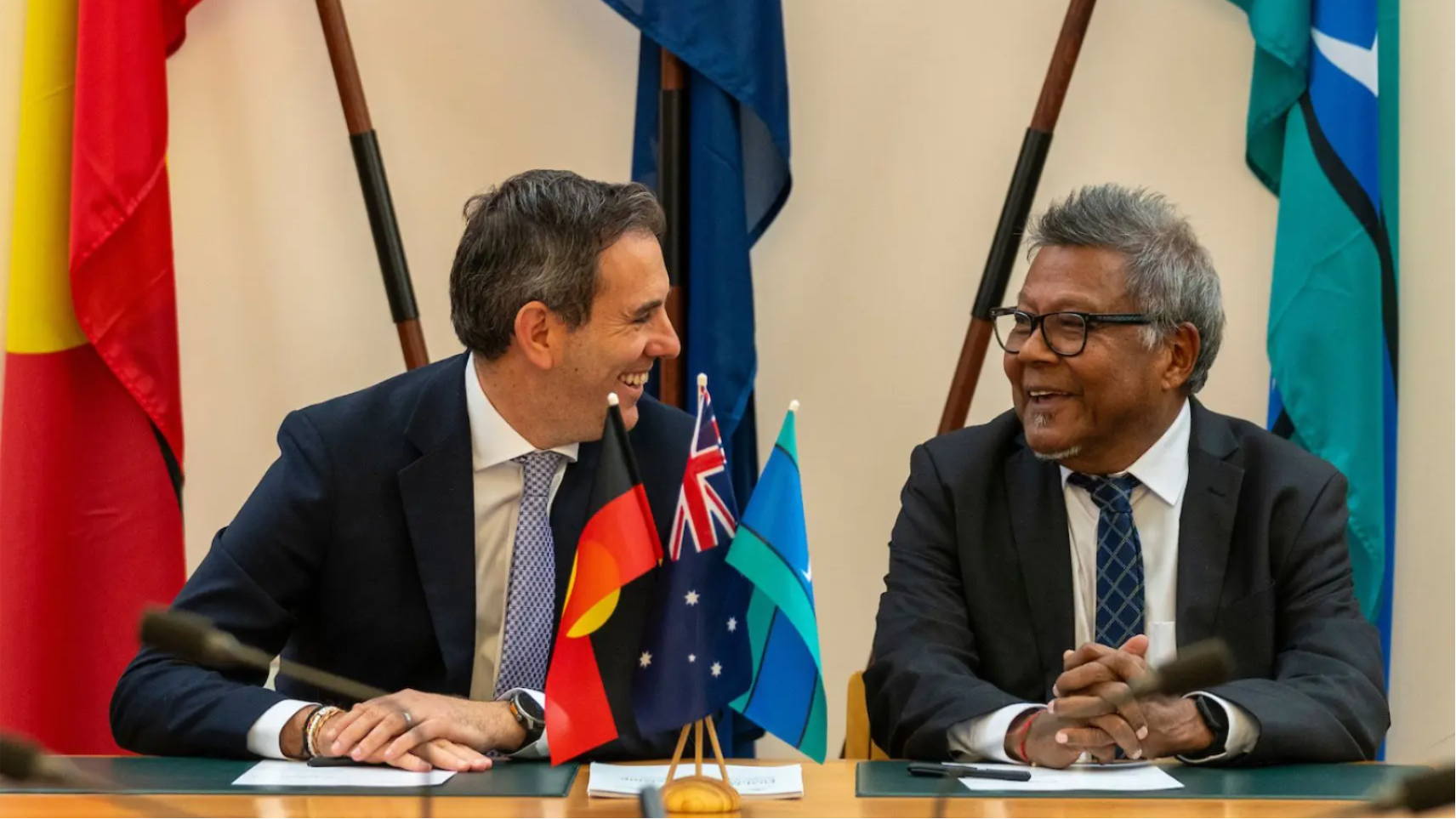 Treasurer Jim Chalmers and Professor Peter Yu smiling in conversation with flags the Aboriginal Flag, Australian flag and Torres Strait Island flags in the foreground and background. 