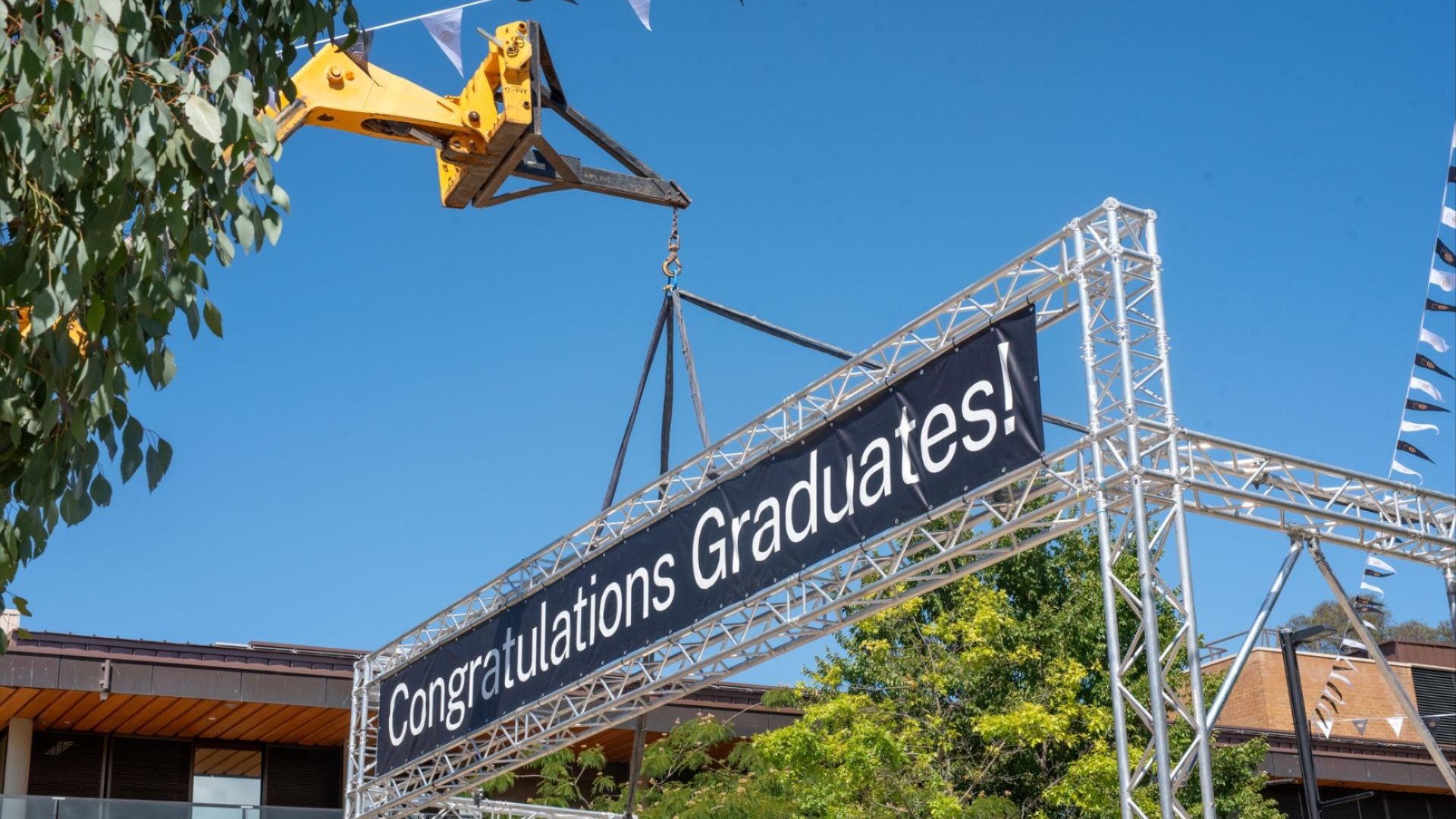 ANU workers use a crane to install a &ldquo;Congratulations Graduates!&rdquo; banner over a campus road.
