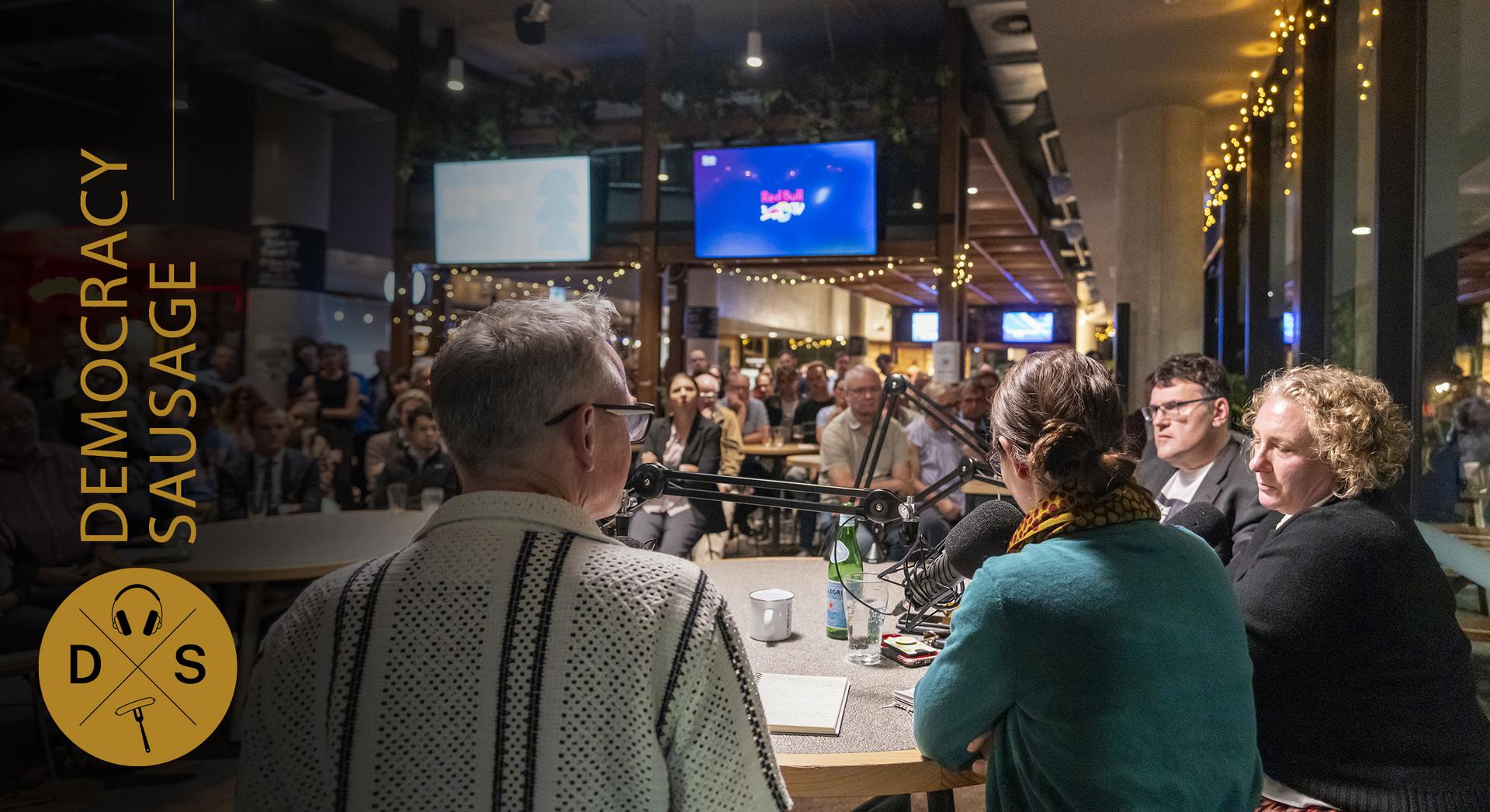Panel discussion recorded live at a venue, with speakers seated at a table facing an audience and microphones, branded Democracy Sausage.
