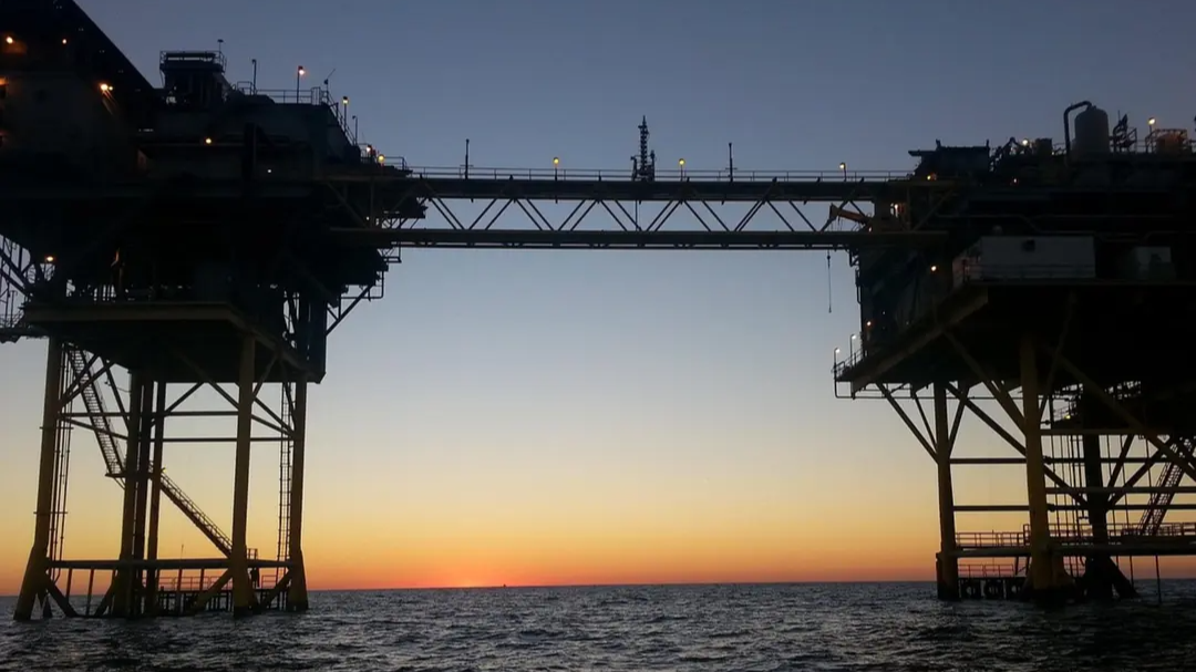 Offshore oil platform silhouetted against a sunset sky, with ocean in the foreground and the sun dipping below the horizon. 