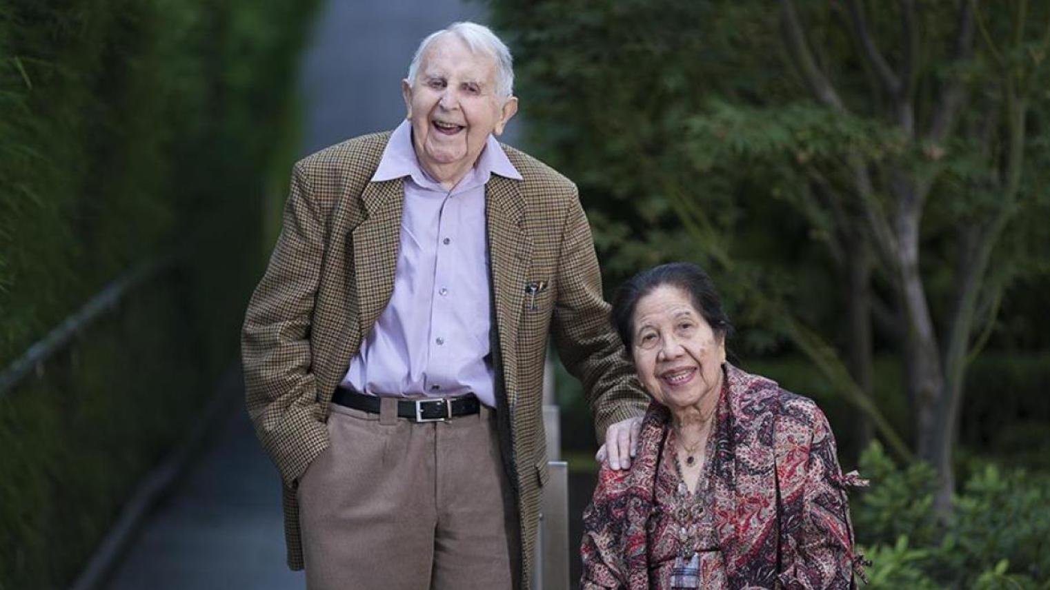 Emeritus Professor Tony Johns and Yohanni Johns smiling outdoors. Tony stands in a brown jacket and the Yohanni is seated in a patterned blouse, with greenery in the background.