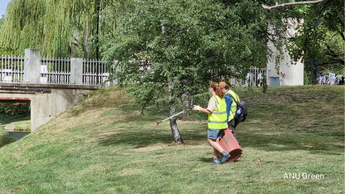 Two people in hi-visibility vests walking on grass by a small river and bridge, carrying a trash bin and litter picker, with trees and a building in the background.