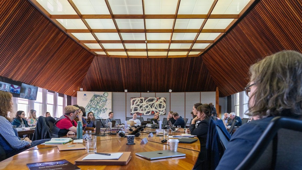 Wide shot of ��������Council members seated around a large table in a timber-lined meeting room, with laptops and papers in front of them.