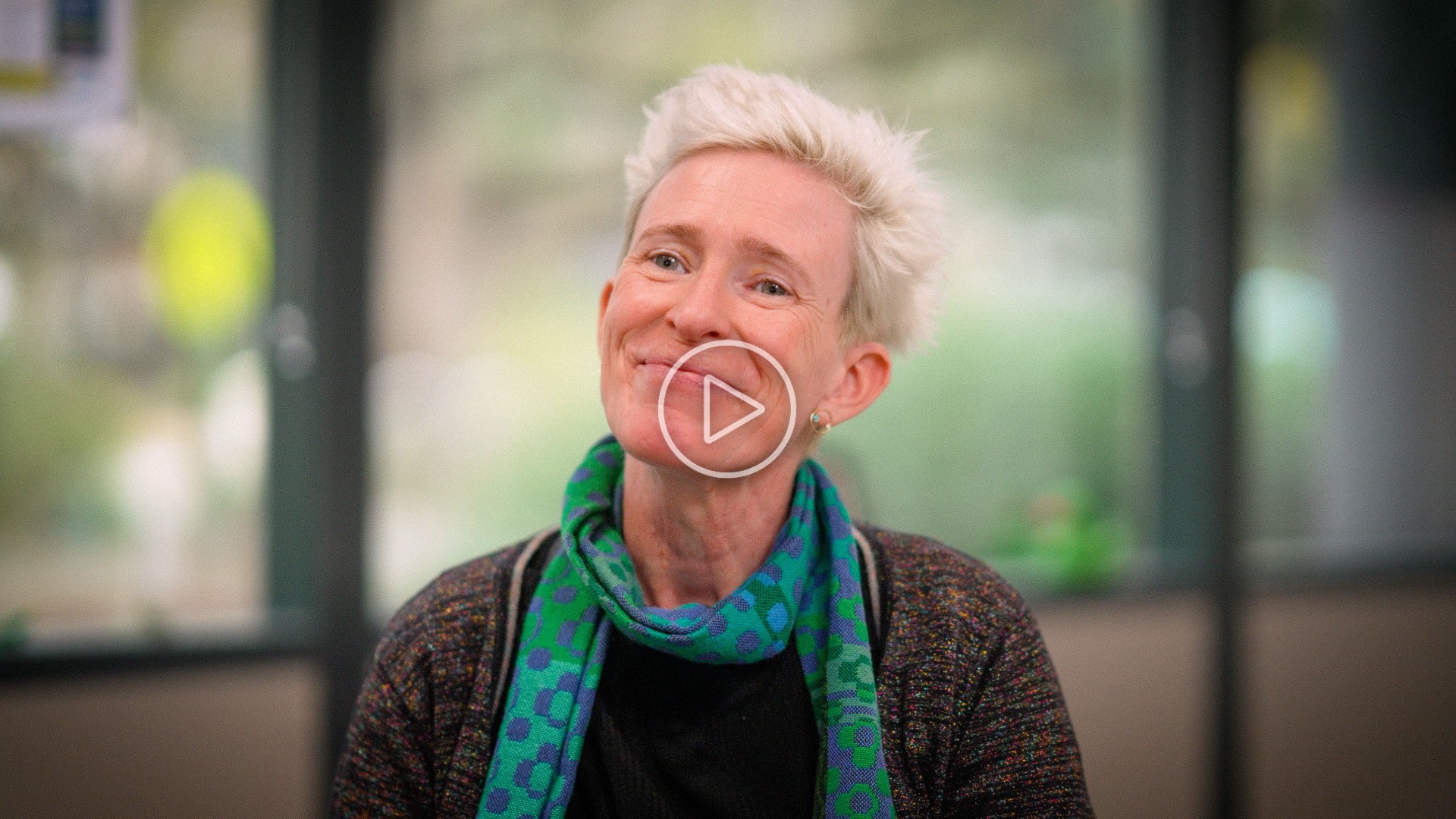 Felicity Gouldthorp smiling indoors, wearing a dark top and a green patterned scarf, with large windows and blurred greenery in the background. 