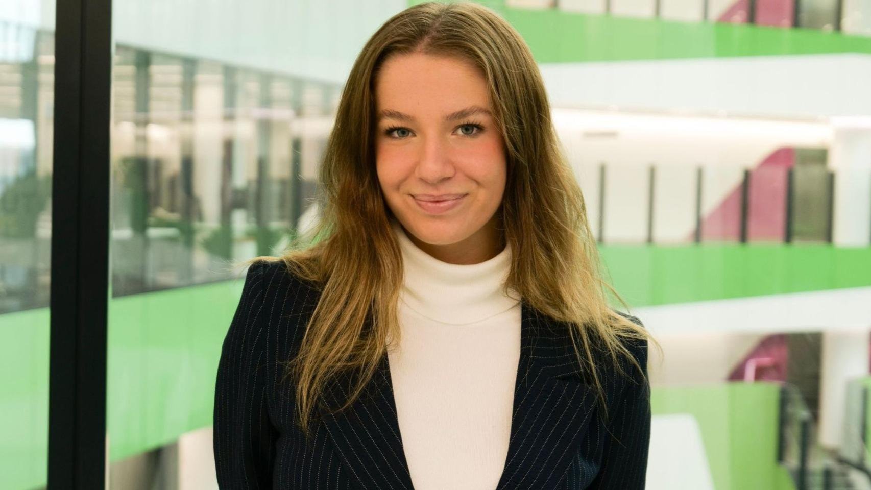 A woman in a dark blazer and white turtleneck smiling indoors with green and white walls in the background.
