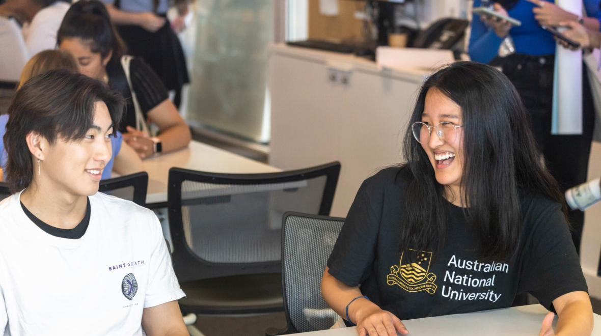 Two ANU students sitting at a table, smiling and laughing during an orientation session.