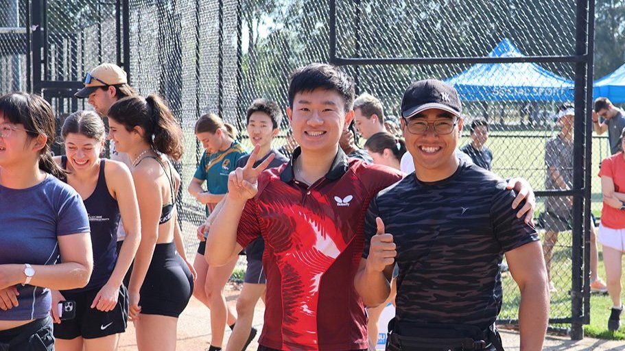 Two students smile and pose with peace signs at an outdoor campus sports event, with others gathered in the background.