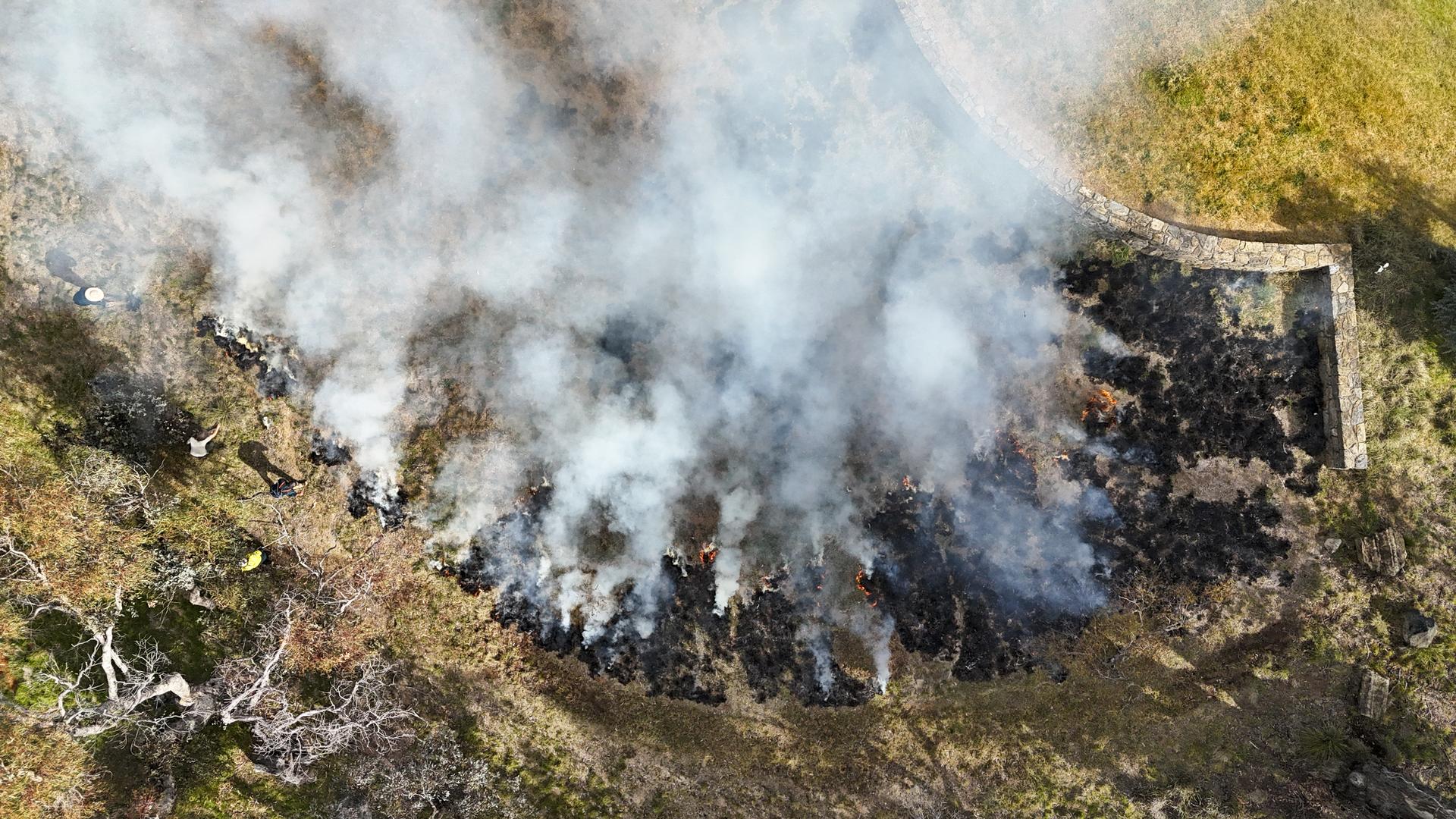 Aerial view of a controlled cultural burn with smoke rising across a grassy area.