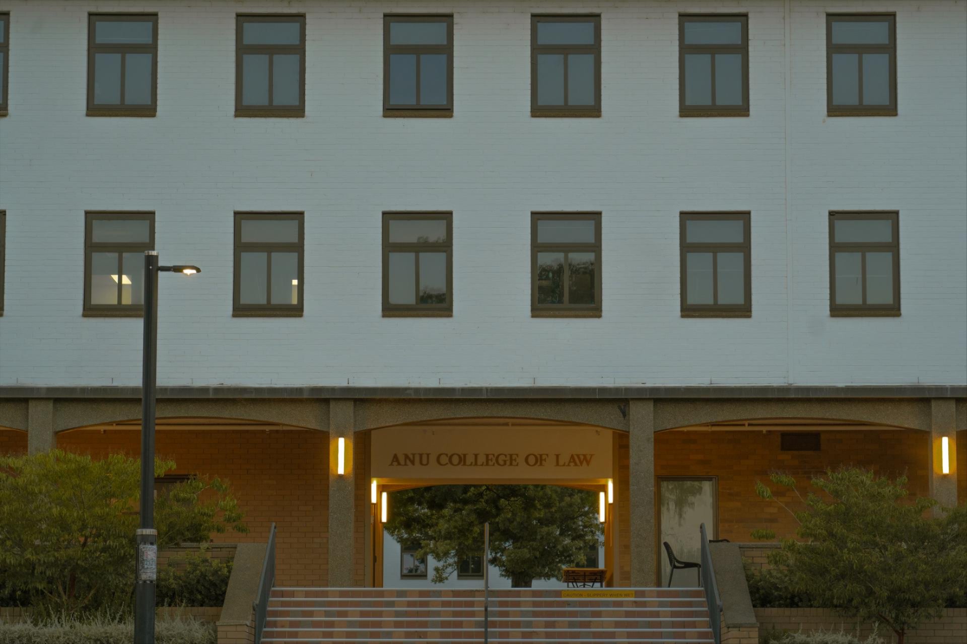 Front entrance of the ANU College of Law building at dusk, with warm lights illuminating the doorway and surrounding trees.