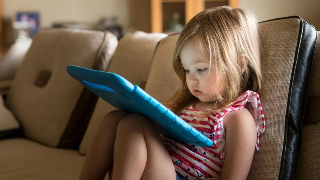 Young child sitting on a couch, focused on a tablet with a blue case.