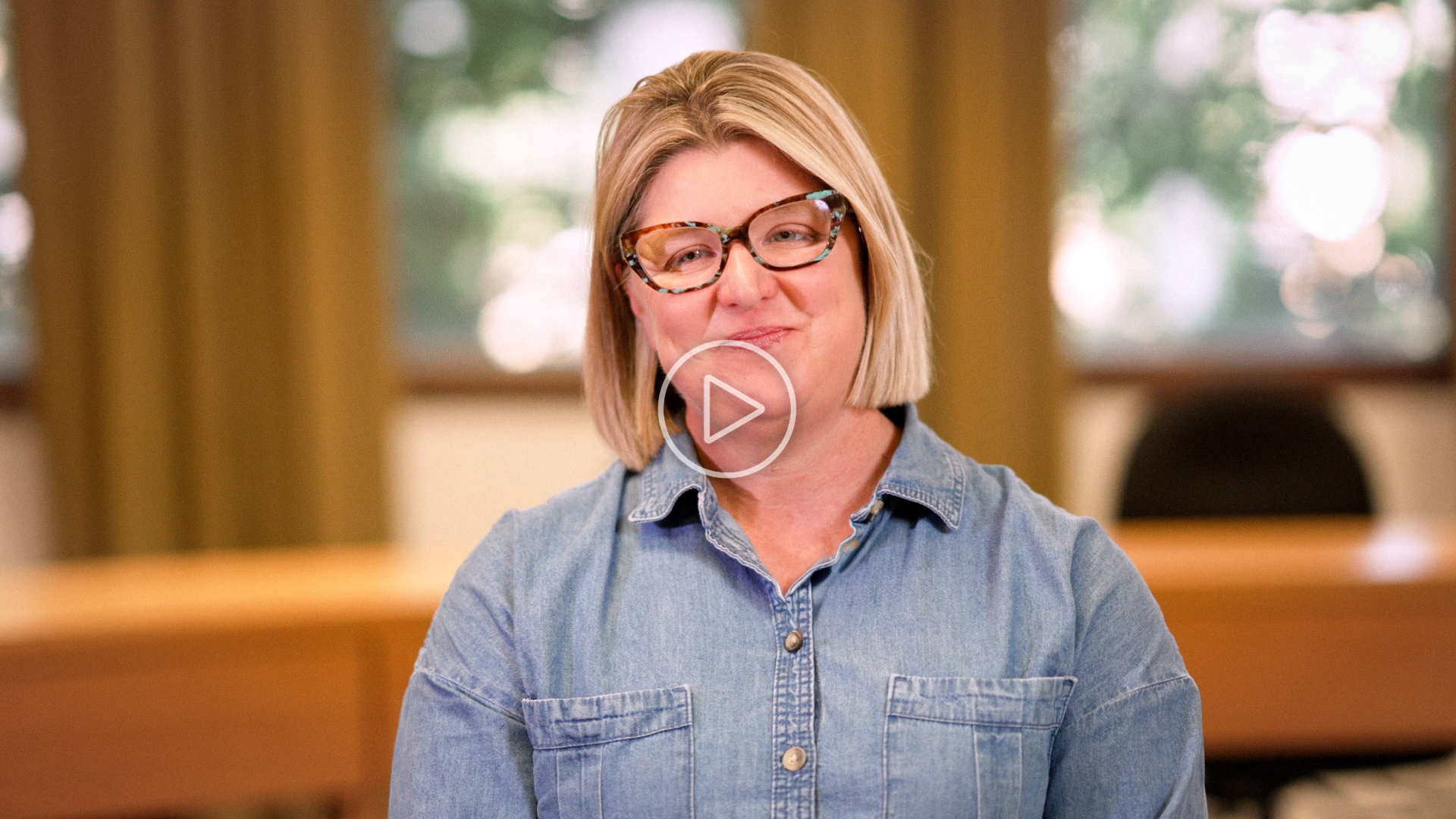Theona Gevorgian smiling indoors, wearing glasses and a light denim shirt, with blurred windows and trees in the background.