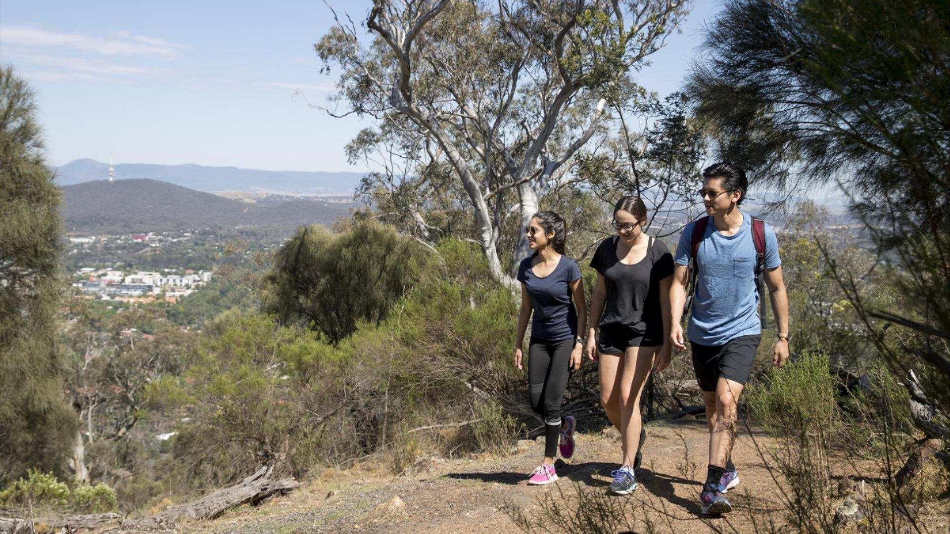 Alexander Longmore smiling while seated outdoors at a table, wearing a dark coat, black shirt, and cap, with trees and umbrellas in the background.