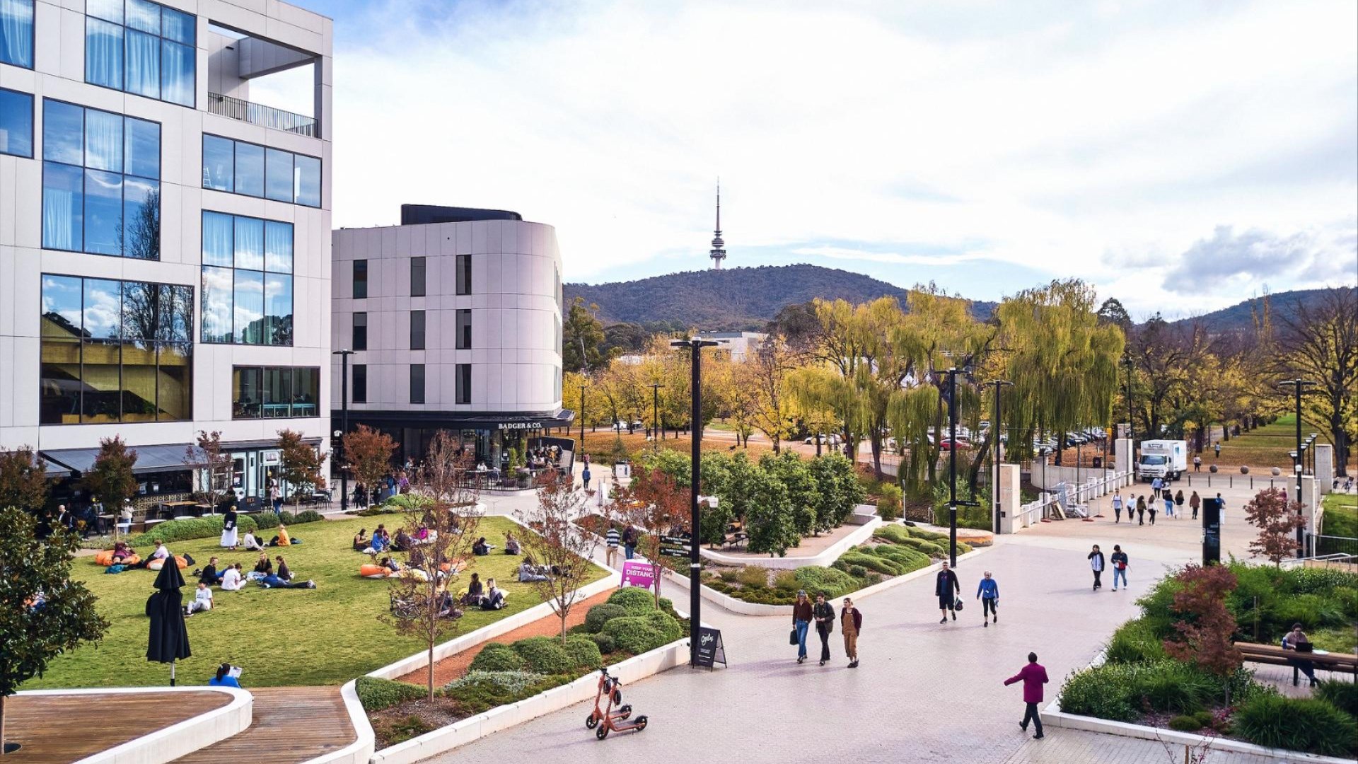 People relaxing on the lawn at Kambri, ANU, with modern buildings and Telstra Tower in the background.
